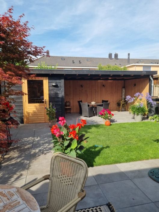 Houten overkapping op maat naast tuinhuis met terras, gazon en bloemen