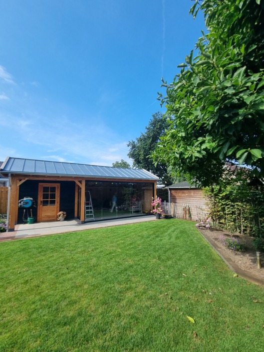 Zwart houten tuinhuis met overkapping aan de rand van een ruime tuin